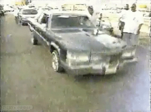 A Man Is Standing Next To A Car That Is Driving Down A Street . GIF