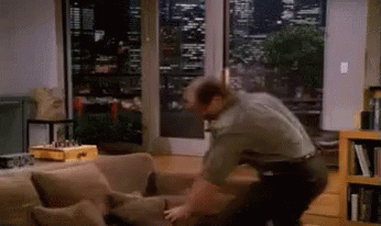 A Man Is Standing Next To A Couch In A Living Room Looking Out A Window GIF