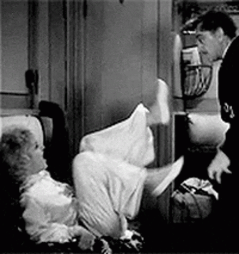A Man Is Standing Next To A Woman Laying On A Couch . GIF