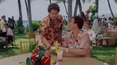 A Man Is Standing Next To A Woman Sitting At A Table With A Drink On It . GIF