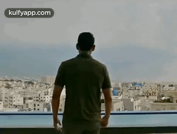 A Man Is Standing On A Balcony Overlooking A City And A Swimming Pool . GIF
