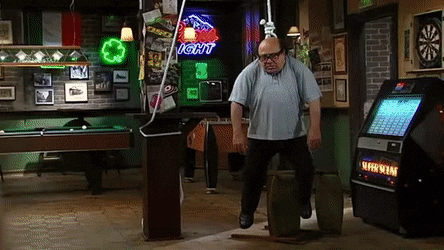 A Man Is Standing On A Barrel In A Bar With A Light Beer Sign Behind Him . GIF