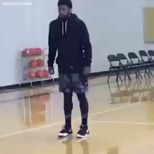 A Man Is Standing On A Basketball Court Wearing A Black Hoodie And Shorts . GIF