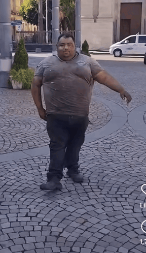 A Man Is Standing On A Cobblestone Street With A White Car Parked Behind Him GIF