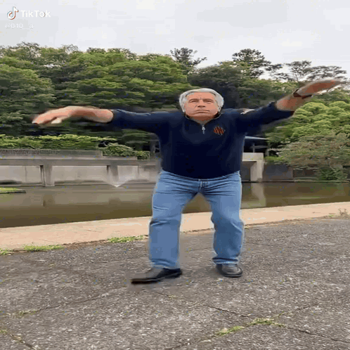 A Man Is Standing On A Sidewalk With His Arms Outstretched In Front Of A River . GIF