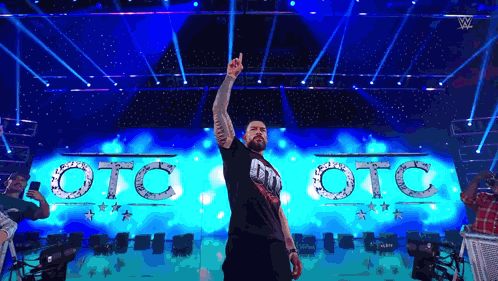 A Man Is Standing On A Stage With His Hand In The Air In Front Of A Sign That Says Oto Oto GIF