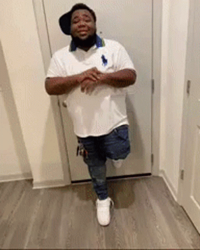A Man Is Standing On One Leg In Front Of A Door Wearing A Hat And A White Shirt . GIF