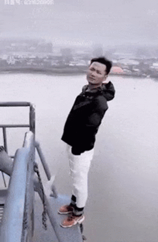 A Man Is Standing On The Edge Of A Bridge Overlooking A Body Of Water . GIF