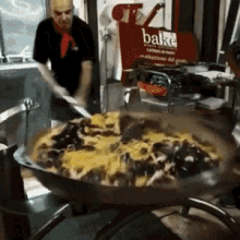 A Man Is Stirring A Large Pan Of Food In Front Of A Sign That Says Bake GIF
