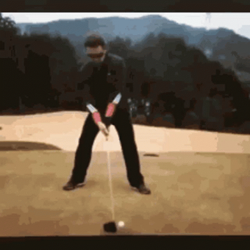 A Man Is Swinging A Golf Club At A Ball On A Golf Course . GIF