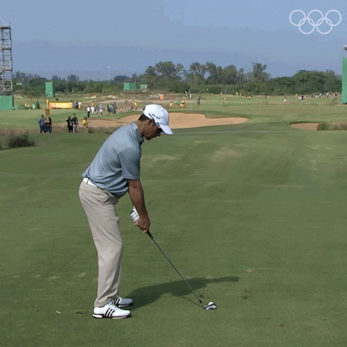 A Man Is Swinging A Golf Club On A Golf Course With The Olympic Rings In The Background GIF