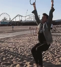 A Man Is Swinging On A Rope Swing On A Beach With A Roller Coaster In The Background GIF