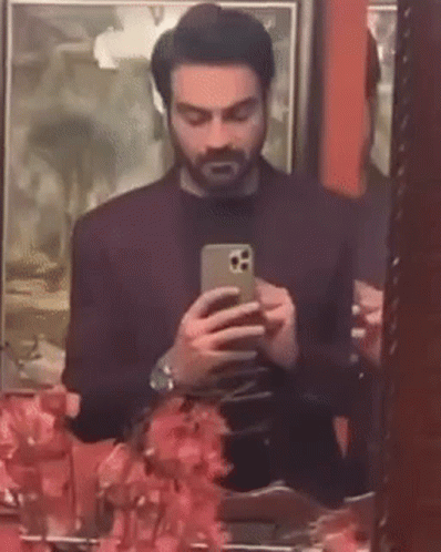 A Man Is Taking A Selfie In Front Of A Mirror While Looking At His Phone . GIF