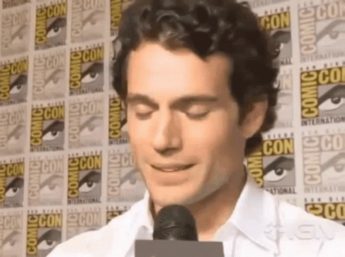 A Man Is Talking Into A Microphone In Front Of A Wall That Says Comic Con GIF