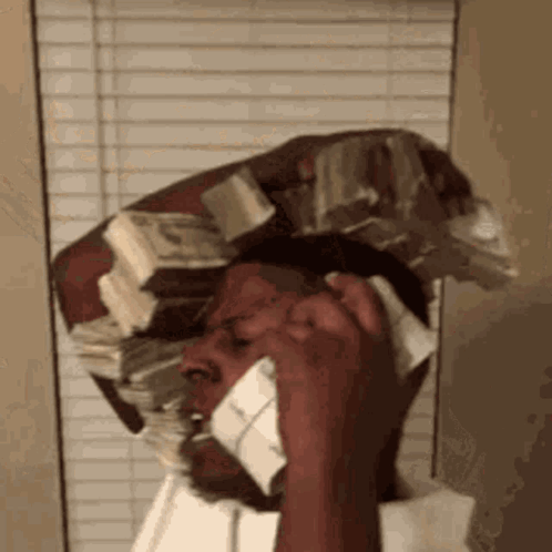 A Man Is Talking On A Cell Phone While Wearing A Hat Made Of Money . GIF