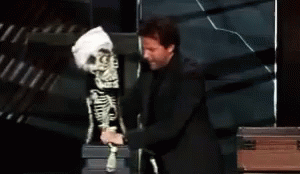 A Man Is Touching A Skeleton Puppet With The Word Touching Behind Him GIF