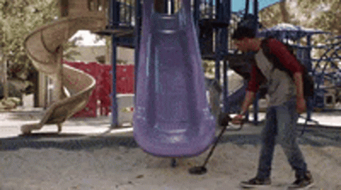A Man Is Using A Metal Detector In A Playground In Front Of A Slide GIF