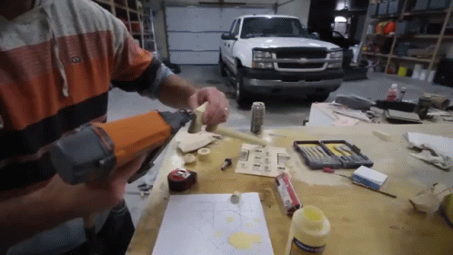 A Man Is Using A Nail Gun On A Piece Of Wood In Front Of A Truck GIF