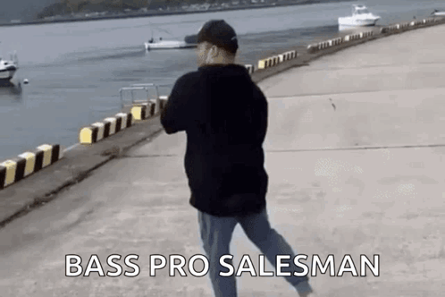 A Man Is Walking Along A Pier Next To A Body Of Water With Boats In The Background . GIF