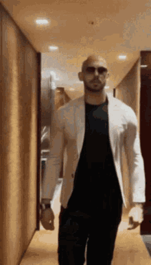 A Man Is Walking Down A Hallway Wearing Sunglasses And A White Jacket . GIF