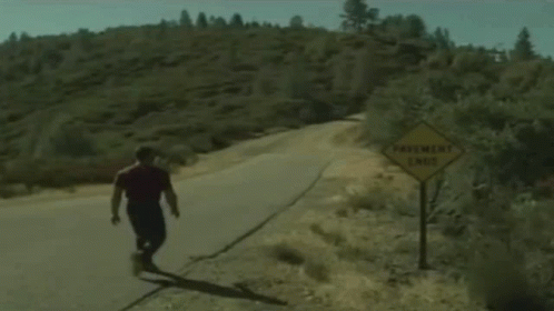 A Man Is Walking Down A Road Next To A Sign That Says Pavement Ends GIF