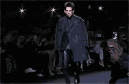 A Man Is Walking Down A Runway Wearing A Black Coat GIF