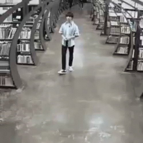 A Man Is Walking Through A Library Filled With Bookshelves . GIF