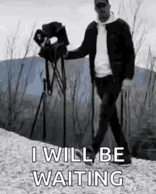 A Man Is Walking With A Camera On A Tripod While Holding A Camera . GIF
