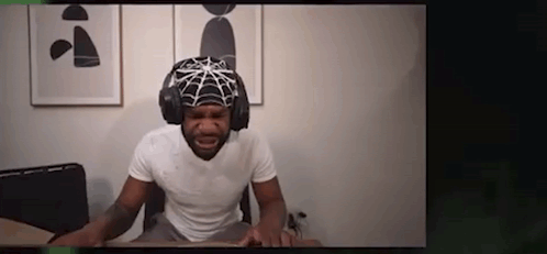 A Man Is Wearing A Spider Web Hat And Headphones While Sitting At A Desk . GIF