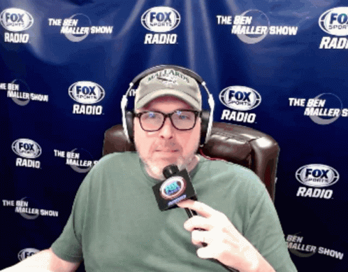 A Man Is Wearing Headphones And Holding A Microphone In Front Of A Fox Sports Radio Banner GIF