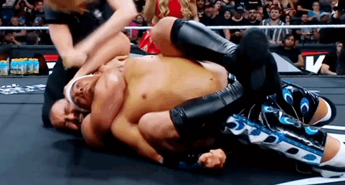 A Man Is Wrestling Another Man In A Wrestling Ring While A Referee Watches . GIF