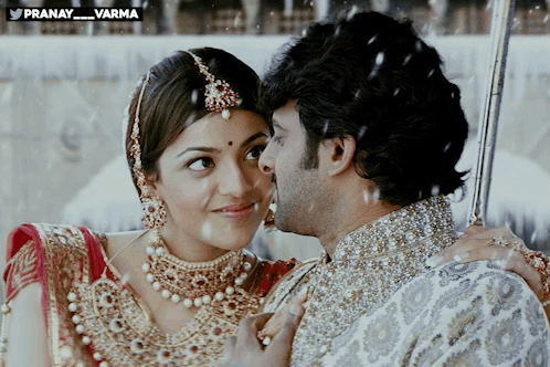 A Man Kisses A Woman On The Cheek In Front Of A Sign That Says Pranay_varma GIF