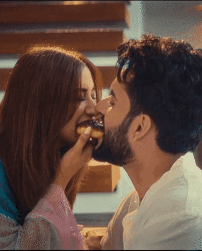 A Man Kisses A Woman On The Cheek While She Eats A Doughnut GIF
