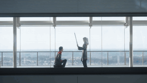 A Man Kneeling Down With A Woman Holding A Stick In Front Of A Large Window GIF