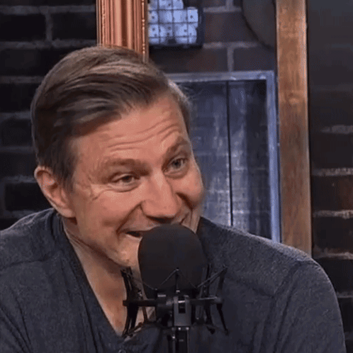 A Man Laughs In Front Of A Microphone With A Brick Wall Behind Him GIF