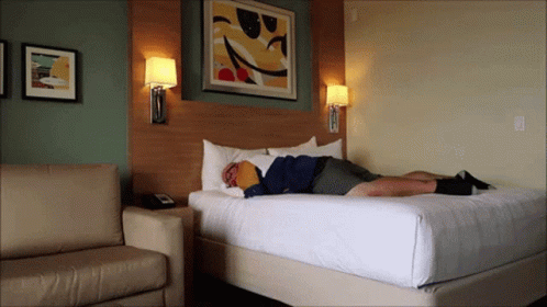 A Man Laying On A Bed In A Hotel Room With A Smiley Face On The Wall GIF