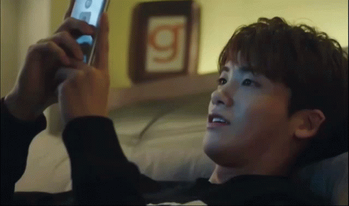 A Man Laying On A Bed Looking At A Cell Phone With The Letter G On The Wall Behind Him GIF