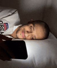 A Man Laying On A Bed Looking At His Phone With A Shirt That Says Brooklyn Great GIF