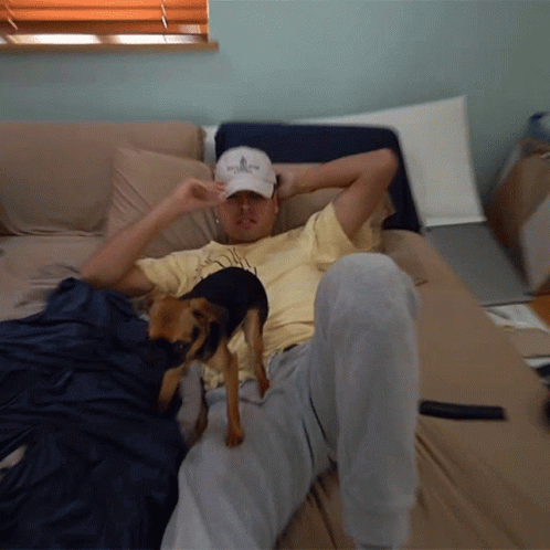 A Man Laying On A Bed With A Dog Wearing A Hat That Says ' Abercrombie ' On It GIF