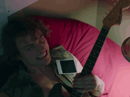 A Man Laying On A Bed With A Guitar And A Cell Phone GIF