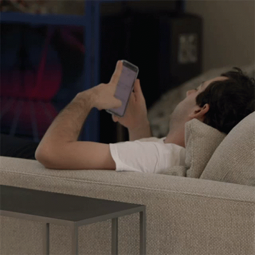 A Man Laying On A Couch Looking At A Cell Phone GIF