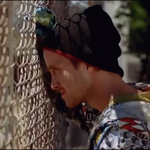 A Man Leaning Against A Chain Link Fence Wearing A Beanie And Sunglasses GIF