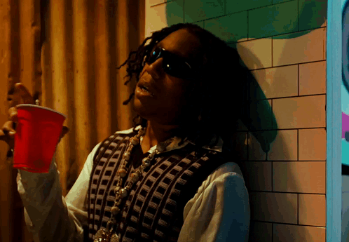 A Man Leaning Against A Wall Holding A Red Plastic Cup GIF