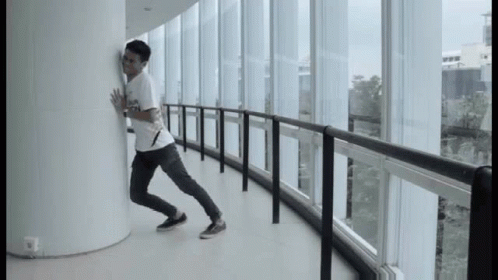 A Man Leaning Against A Wall In A Hallway With Lots Of Windows GIF