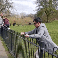 A Man Leaning Over A Fence In A Park With A Woman Sitting Behind Him GIF
