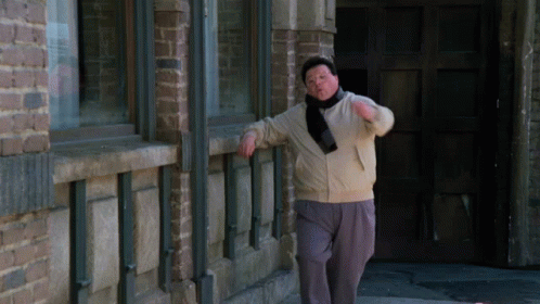 A Man Leans Against A Brick Wall While Wearing A Scarf Around His Neck GIF