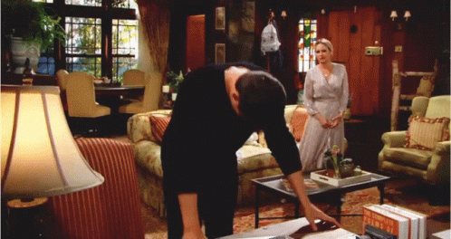 A Man Leans Over A Table In A Living Room With A Woman Standing In The Background GIF