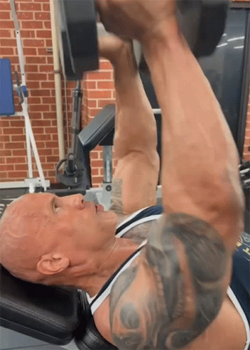 A Man Lifting A Dumbbell In A Gym With A Brick Wall In The Background GIF