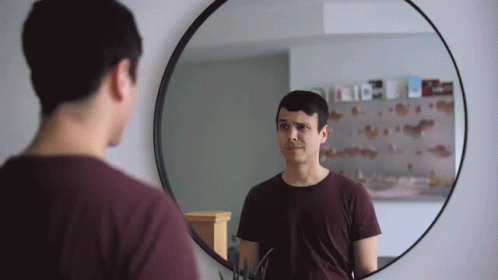 A Man Looks At Himself In A Round Mirror With A Reflection Of A Shelf With Books On It GIF