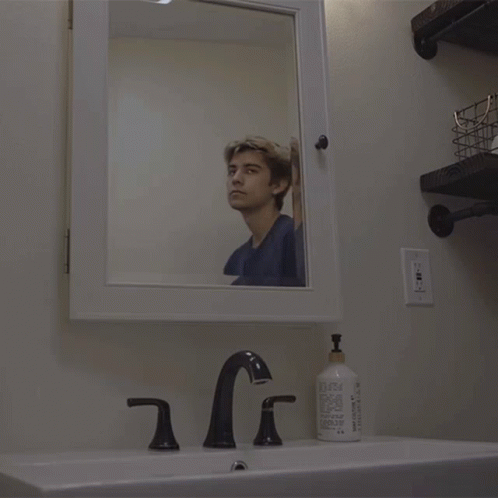 A Man Looks At His Reflection In A Mirror In A Bathroom GIF